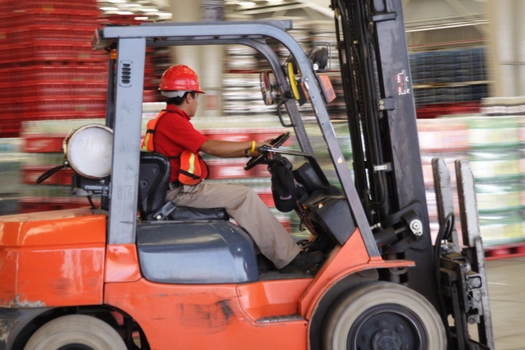 A young worker in a red shirt and helmet drives a forklift through a warehouse filled with stacked crates