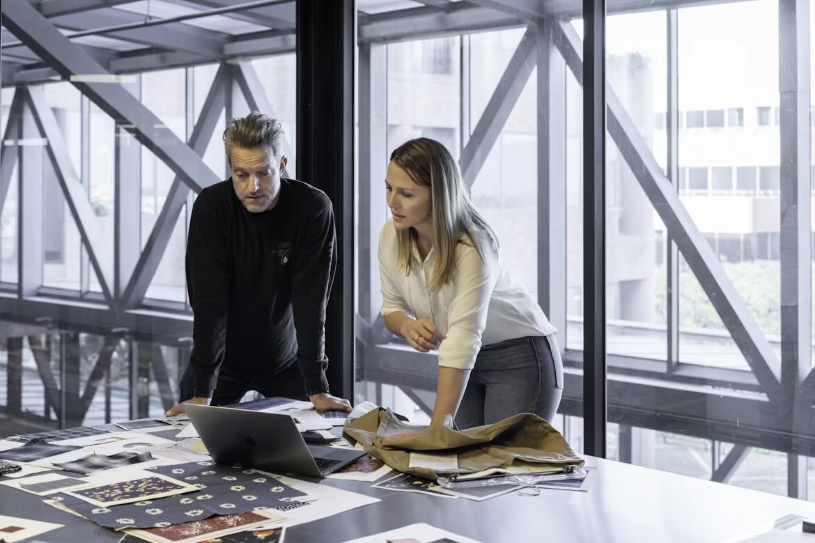 Two young professionals standing over a laptop and a table covered in sample pages